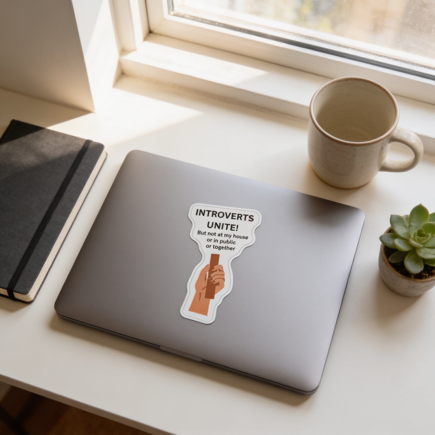 a laptop with a sticker on it, a notebook, a coffee cup, and a potted plant on a desk.